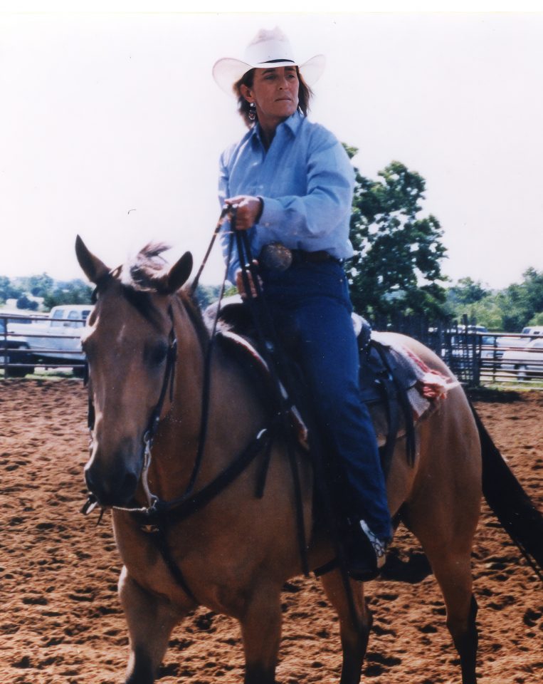 Person wearing a white cowboy hat and blue shirt rides a brown horse in a dusty outdoor arena.