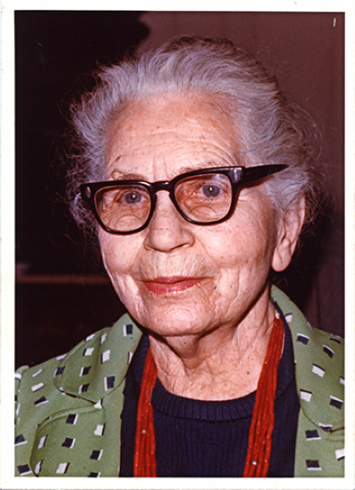 Portrait of an elderly woman with white hair and black-frame glasses, wearing a green patterned jacket over a navy top with a red scarf.