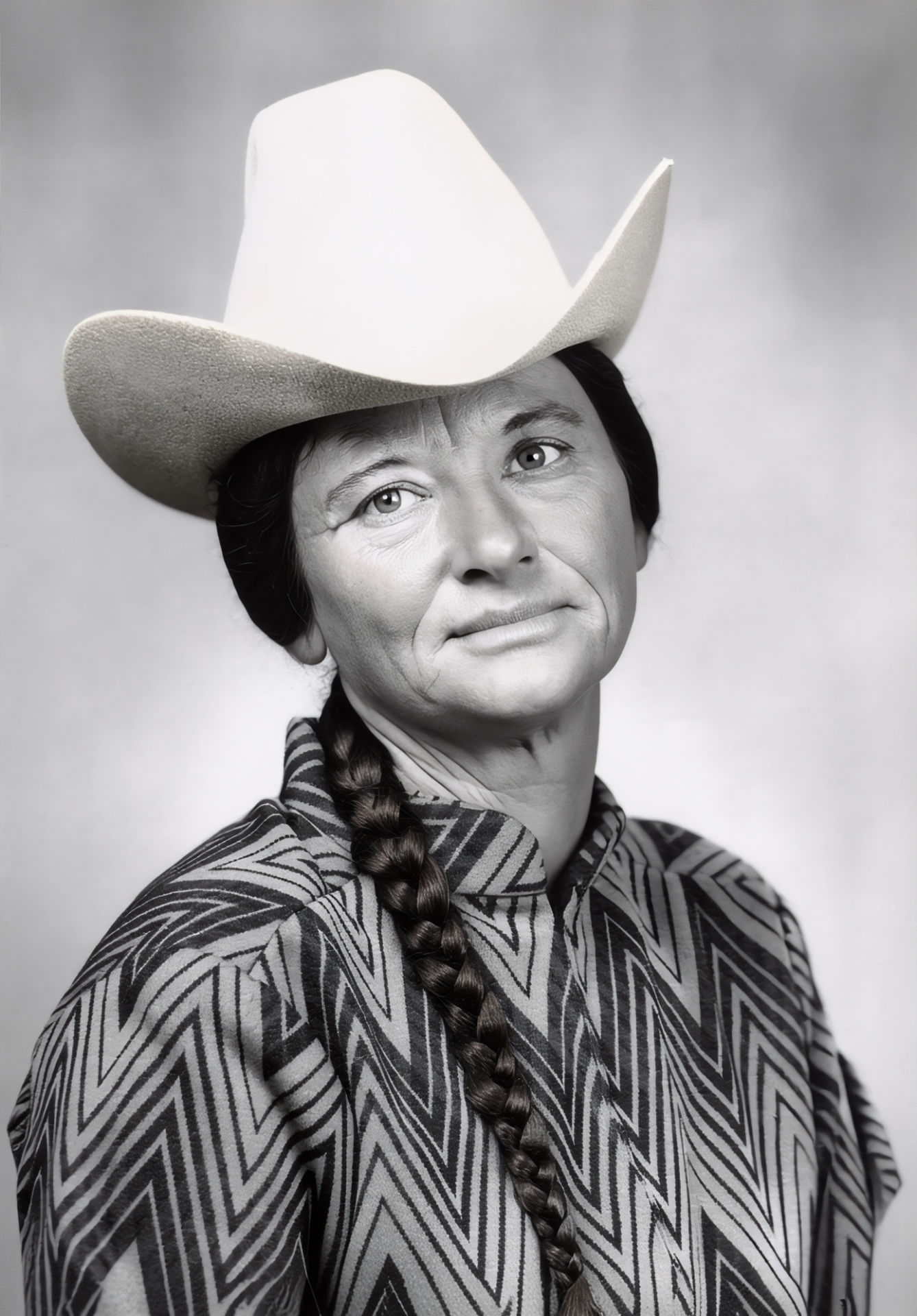 Portrait of a woman in a large white cowboy hat, braided hair, and a zebra-like patterned jacket, looking softly at the camera.