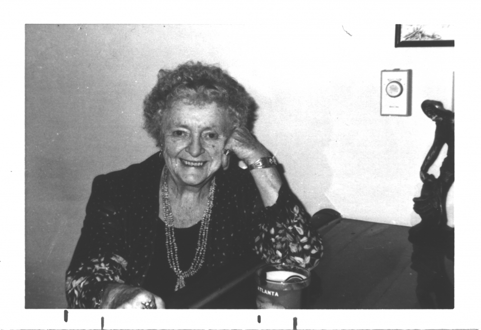 Smiling elderly woman with short curly hair, wearing beaded necklaces, sits at a table indoors.
