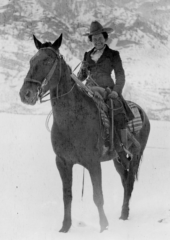 Woman in a wide-brimmed hat riding a dark horse through a snowy, mountainous landscape; both rider and horse are equipped with western tack.
