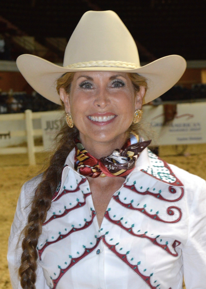Smiling woman in a white cowboy hat with braided hair, wearing a patterned western shirt and scarf.