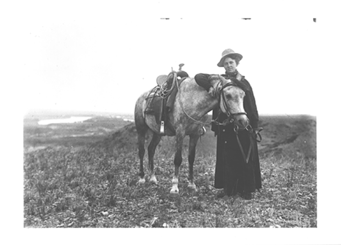 Woman standing beside a saddled horse in a wide, grassy field in a rural landscape with a hat and long coat.