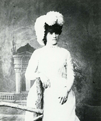 19th-century woman in a long white dress with a feathered hat, standing beside a wooden railing against a studio backdrop.