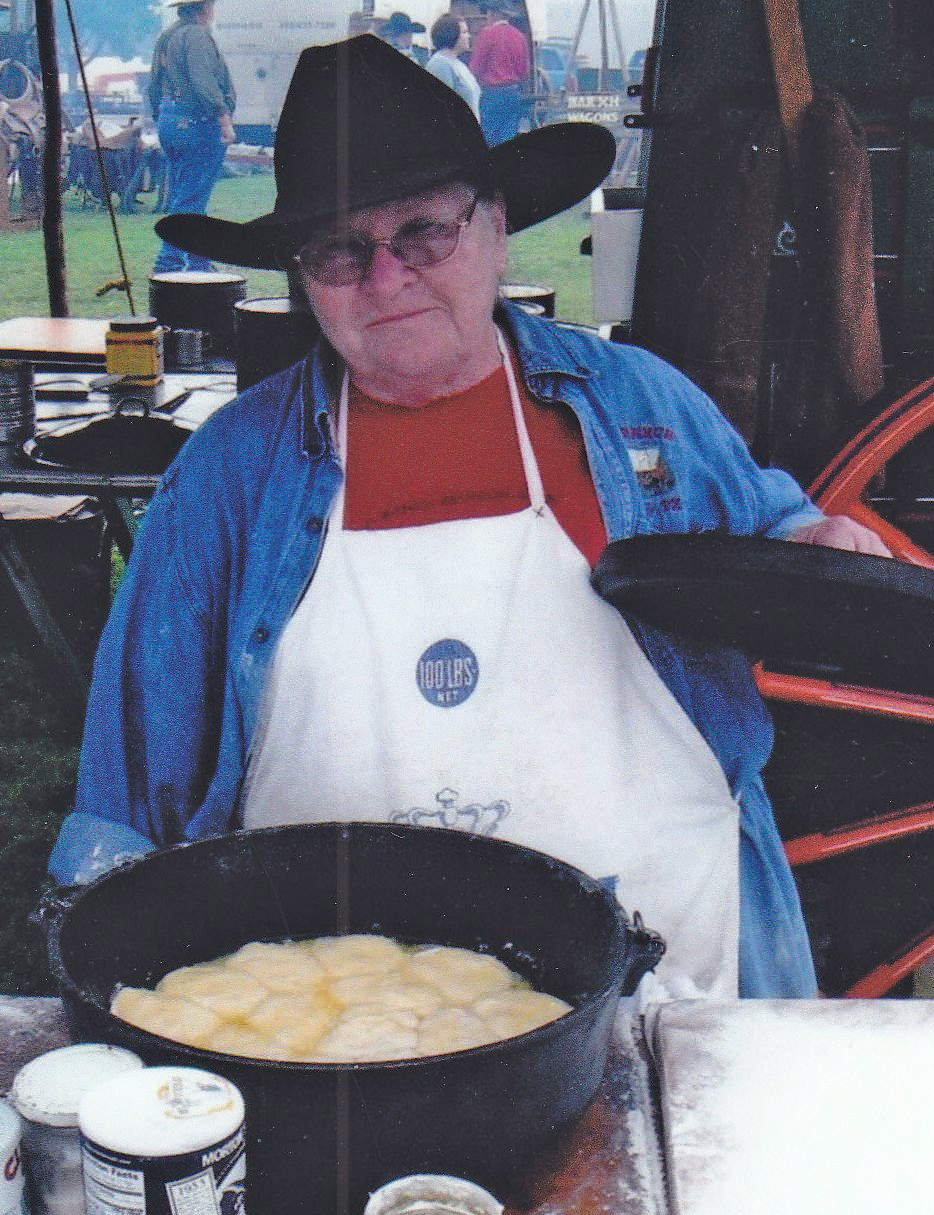 Older man in a black cowboy hat and denim shirt cooking dough in a large cast-iron pan at an outdoor fair.