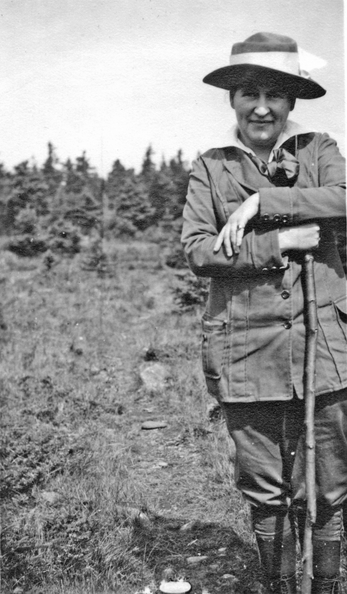 Black-and-white photo of a man in a wide-brimmed hat and field jacket, leaning on a rifle outdoors with trees in the background.