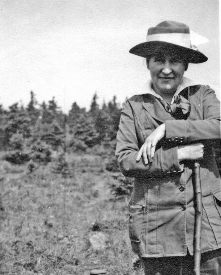 Black-and-white photo of a man in a wide-brimmed hat and field jacket, leaning on a rifle outdoors with trees in the background.
