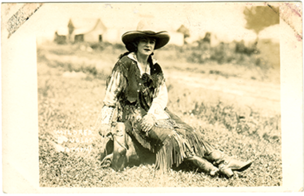 Vintage sepia photo of a woman in a fringed Western dress and wide-brim hat, sitting on grass in an open field.