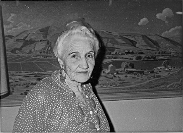 Elderly woman with white hair in a polka dot dress standing in front of a landscape mural, looking at the camera with a subtle smile.