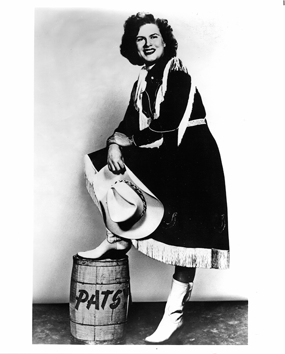 Smiling woman in fringed dress and white boots, leaning on a hat while one foot rests on a barrel labeled 'PATS'.