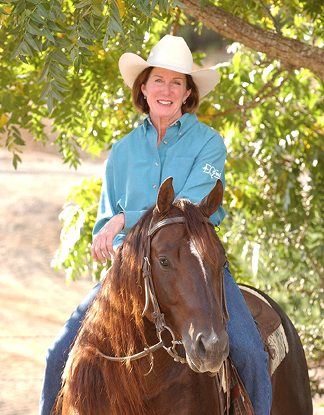 Smiling woman in a white cowboy hat rides a brown horse under a leafy tree.