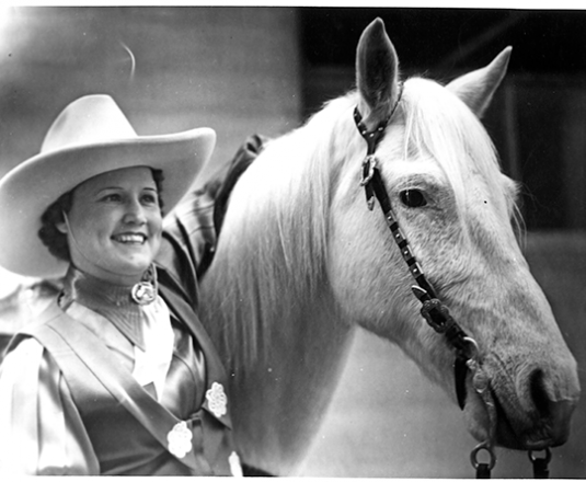 Smiling cowgirl in a wide-brimmed hat beside a white horse with a bridle, facing left.