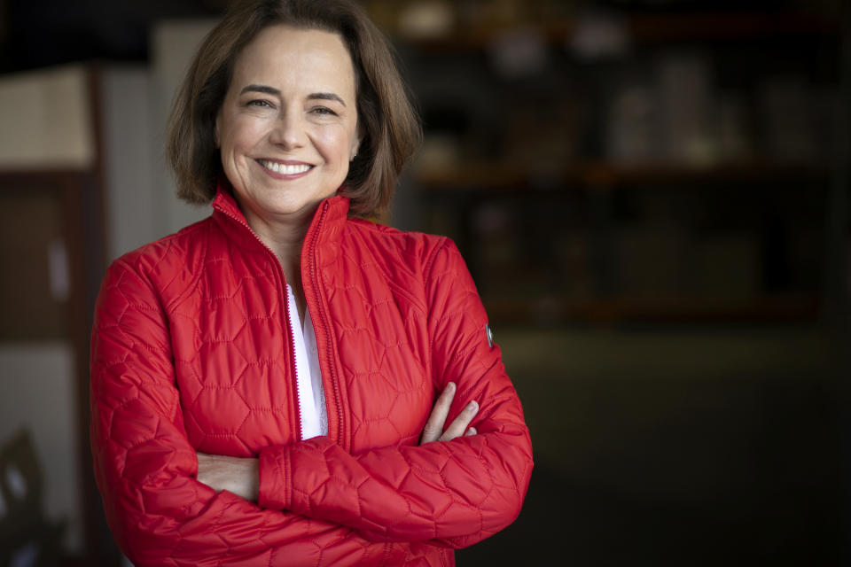 smiling woman in a red quilted jacket with arms crossed, looking at the camera in a casual indoor setting