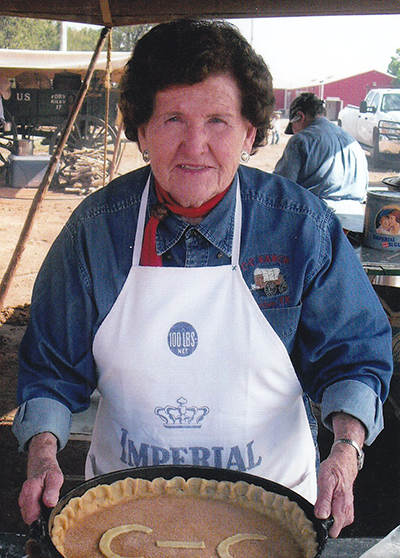 Older woman in a denim shirt and white apron stands behind a large pie in a pan outdoors, the crust decorated with the letters 'C-C' at a fair or festival.