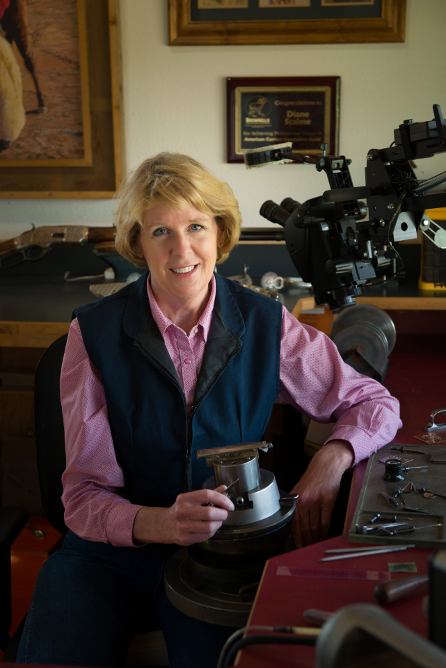Smiling woman in a pink shirt and dark vest sits at a workbench in a workshop, using a metal lathe to shape a part at her desk