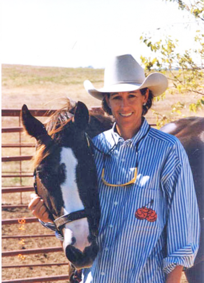 Woman in a white cowboy hat standing beside a horse in a fenced outdoor area; she wears a blue striped shirt and smiles at the camera.