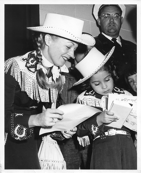 Two young women in Western outfits with fringed jackets and white cowboy hats reading a letter together at a crowd-filled event.