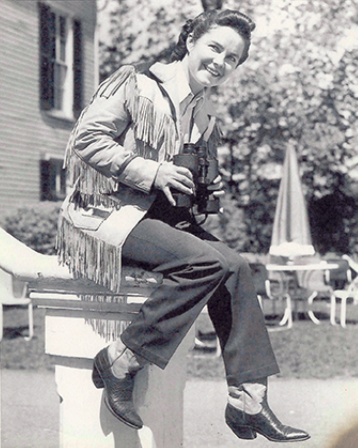 Woman in a fringed jacket sits on a fence outside, smiling and holding binoculars.