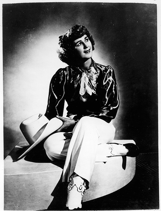 Portrait of a smiling woman in a satin blouse and white pants, seated on a curved stool, holding a wide-brim hat in her lap.