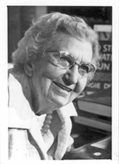 Smiling elderly woman with short curly hair and glasses, wearing a pearl necklace and white blouse.