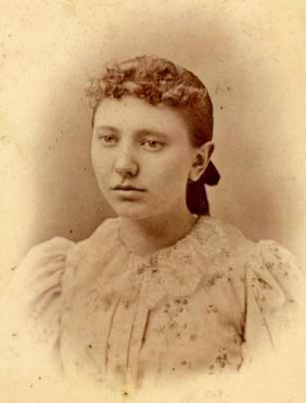 Sepia portrait of a young woman with a bow in her hair, wearing a floral-patterned dress with puffed sleeves.