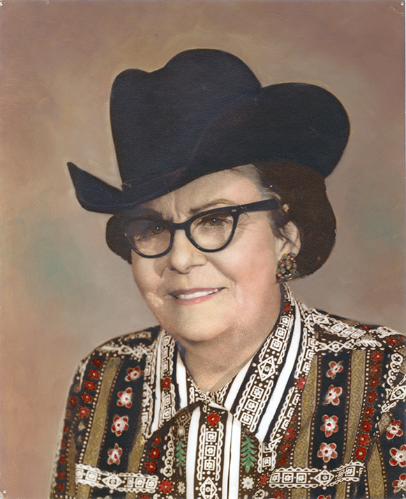 Portrait of an older woman wearing a large black hat and black-framed glasses, smiling, in a colorful patterned blouse and earrings.