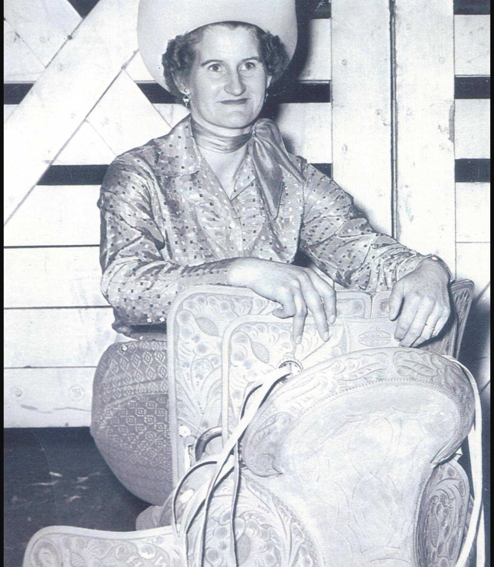 Black-and-white photo of a woman in a wide-brim hat, patterned blouse and scarf, seated on an ornate carved chair.