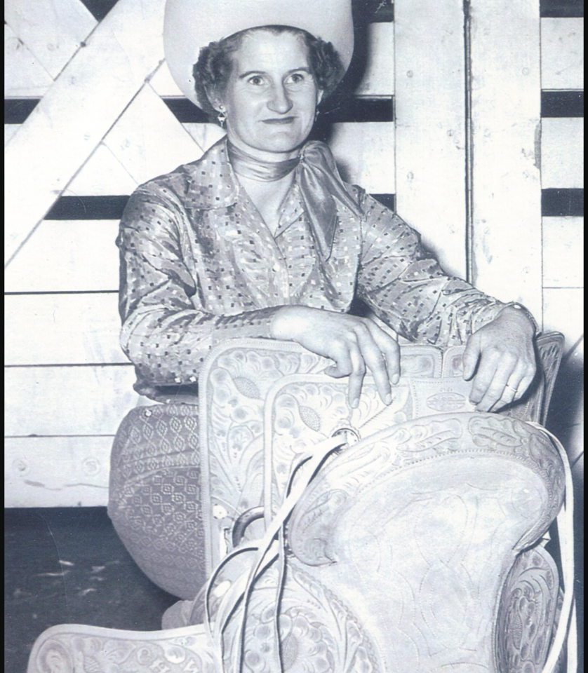 Black-and-white photo of a woman in a wide-brim hat, patterned blouse and scarf, seated on an ornate carved chair.