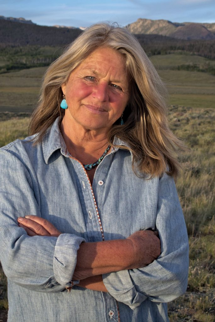 Older woman with long gray-blonde hair wearing a light denim shirt, arms crossed, standing in a grassy field with mountains in the background. - or - Portrait of a woman outdoors, arms crossed, turquoise jewelry, in a rural landscape with hills behind her.