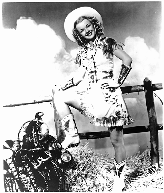 Vintage photo of a smiling cowgirl in a fringed dress and hat, posing with one boot on hay beside a wooden fence.