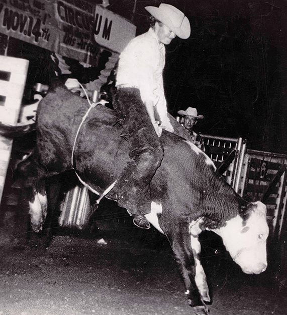 Cowboy in a white shirt and hat rides a bucking bull at a nighttime rodeo arena.
