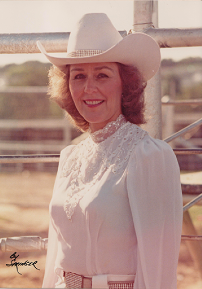 Portrait of a smiling woman in a white lace blouse and wide-brimmed cowboy hat, outdoors by a fence at a ranch.