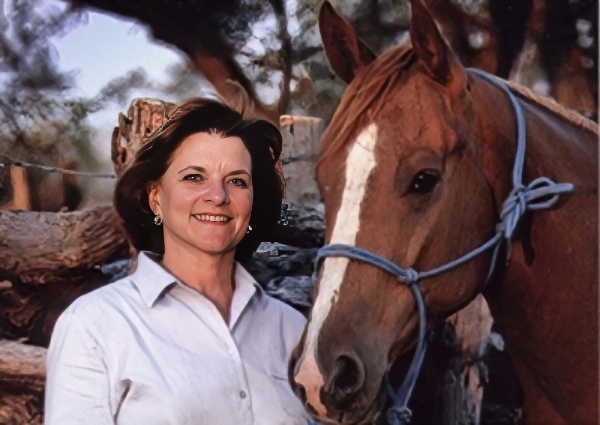 Smiling woman in a white shirt standing beside a brown horse with a blue halter in a rustic outdoor setting.