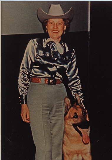 Smiling woman in a silver Western shirt, light gray pants, and a cowboy hat standing next to a large German Shepherd dog in a dark studio setting.