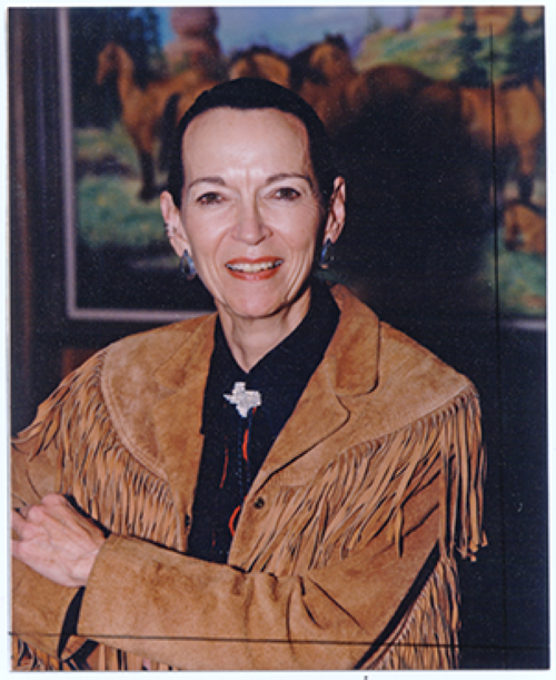 Portrait of a smiling woman wearing a tan suede fringe jacket and black blouse, standing indoors with a painting in the background.