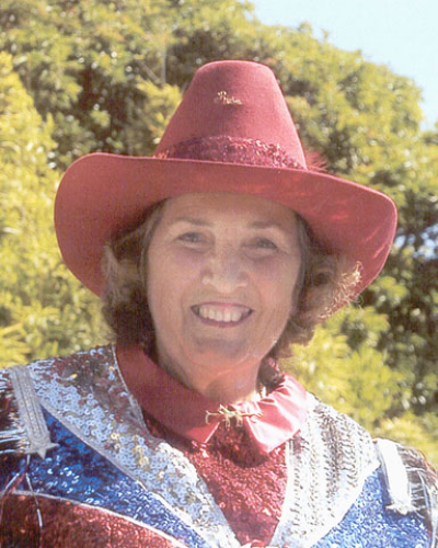 Smiling woman wearing a red wide-brim hat and a sequined jacket, outdoors with green trees in the background.