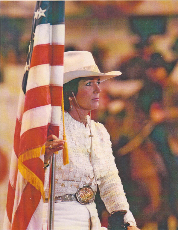 Woman in a white sequined outfit and cowboy hat holding an American flag at a parade or ceremony