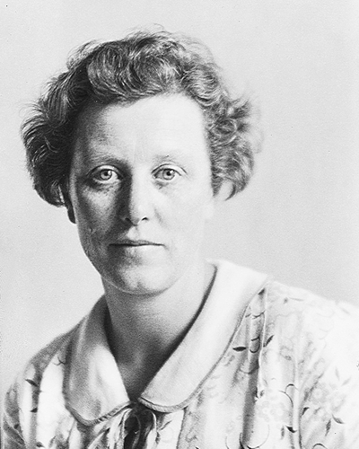 Black-and-white portrait of a woman with short, curly hair and a neutral expression, wearing a patterned blouse with a rounded collar.