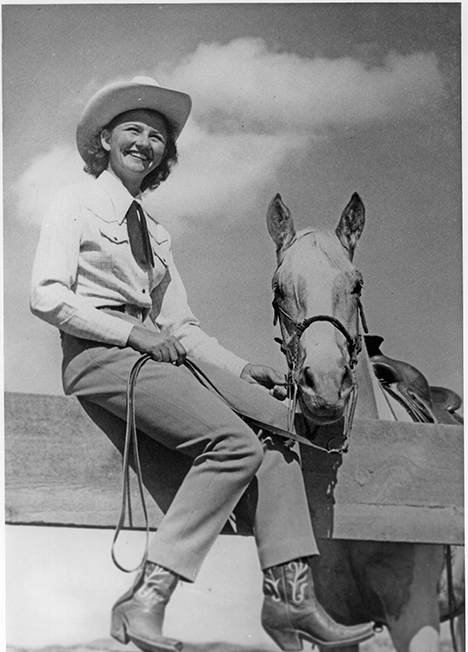 Smiling woman in a cowboy hat sits on a horse, holding the reins outdoors on a sunny day (vintage photo).