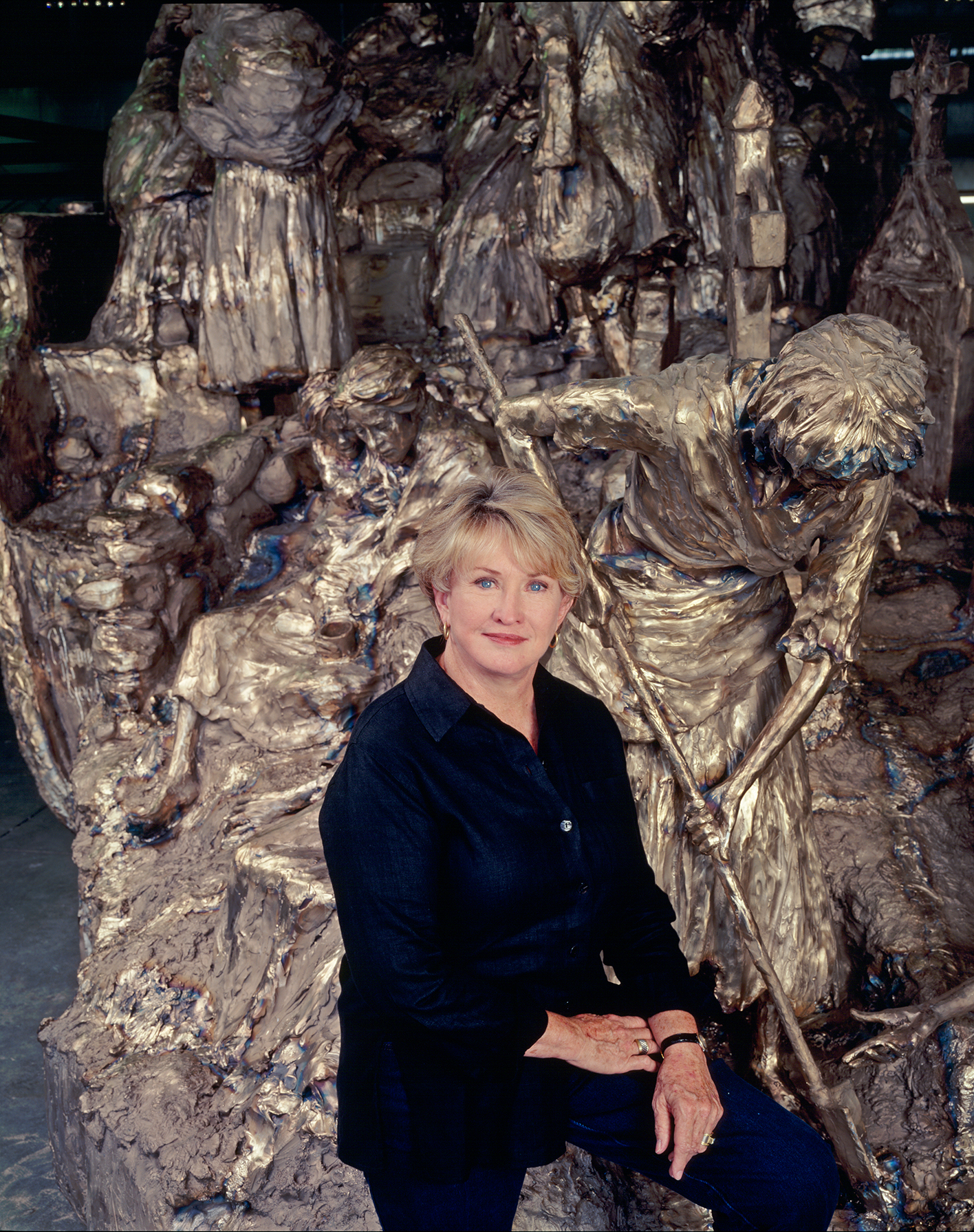 Portrait of a woman with short blond hair wearing a dark shirt, posing in front of a large metallic sculpture resembling melted figures at an art installation.