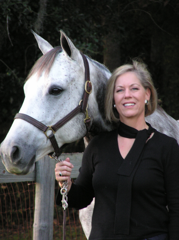 Smiling woman in a black top stands beside a gray horse, holding its lead near a wooden fence.