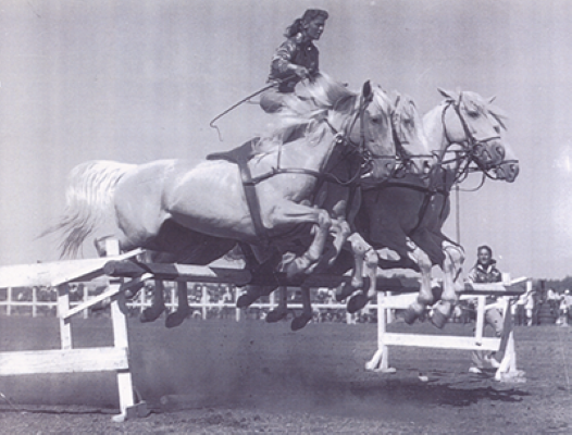Black-and-white photo of a rider and horse mid-jump over a hurdle in a show jumping event, with another horse nearby and spectators in the background