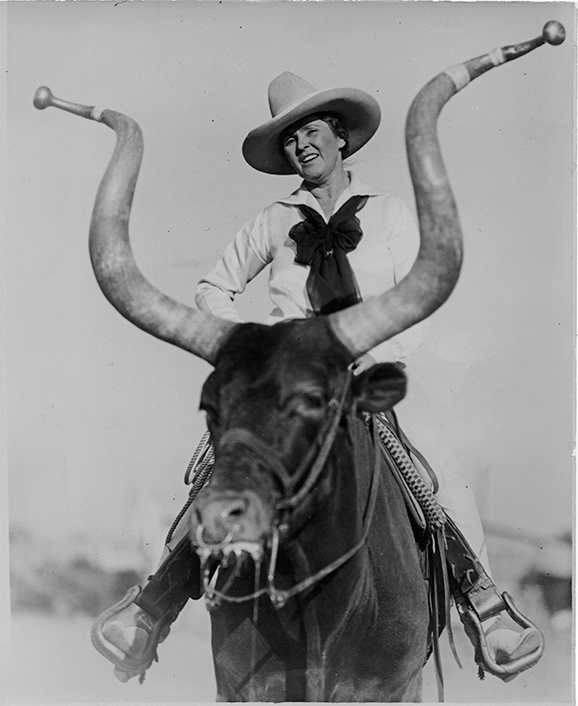 Rider in a cowboy hat and scarf rides a bull with long curved horns, smiling during a rodeo moment.