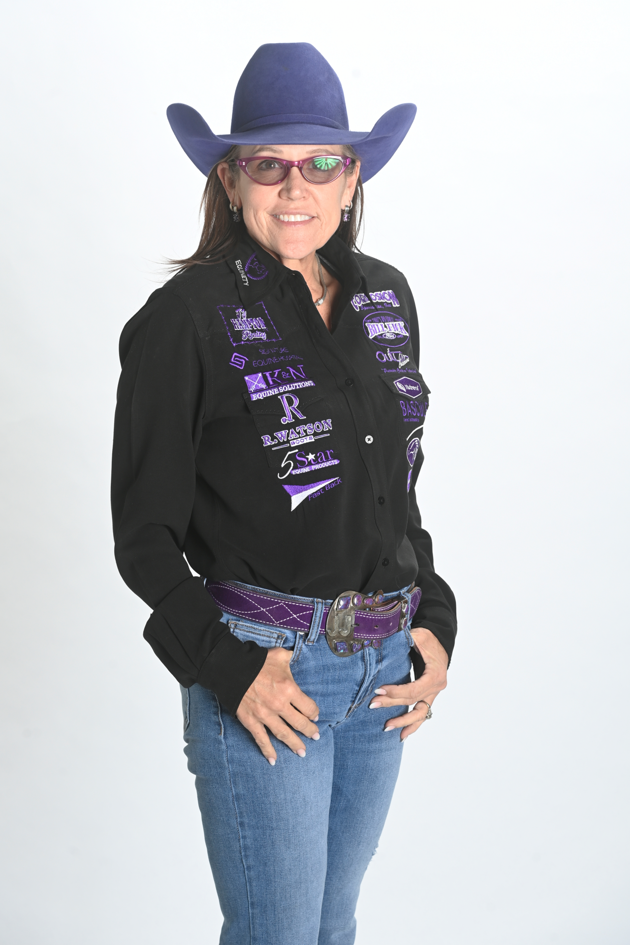 Smiling woman in a purple cowboy hat, glasses, and a black shirt decorated with various colorful embroidered patches, standing against a plain white background.