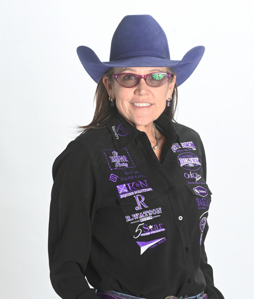 Smiling woman in a purple cowboy hat, glasses, and a black shirt decorated with various colorful embroidered patches, standing against a plain white background.