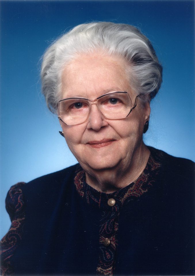 Portrait of an elderly woman with white hair and glasses wearing a dark vest against a blue gradient background, looking at the camera with a gentle smile.
