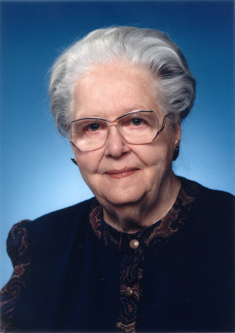Portrait of an elderly woman with white hair and glasses wearing a dark vest against a blue gradient background, looking at the camera with a gentle smile.