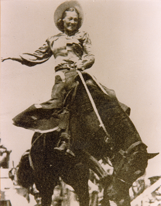 Woman in period dress riding a galloping horse, waving an arm as a crowd watches.