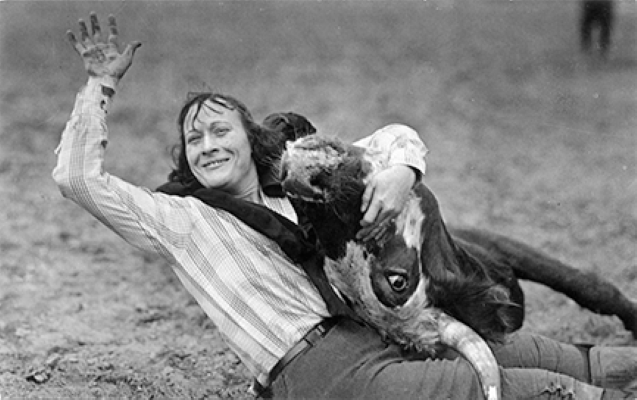 Person lying on the ground in a striped shirt, smiling and waving, as a large dog cuddles against them and rests its head on their arm.