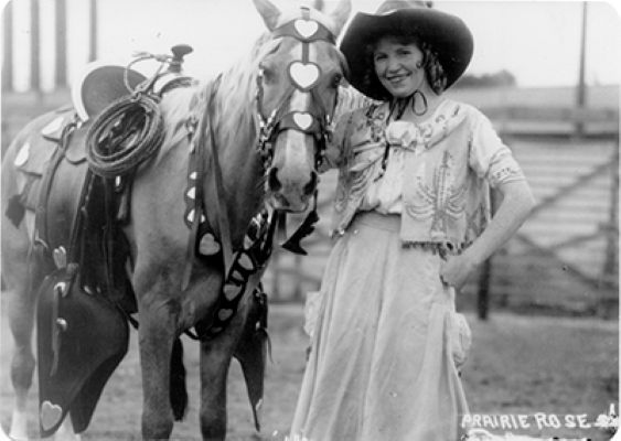 Woman in a wide-brimmed hat and fringed dress stands beside a horse wearing heart-shaped tack in a rural arena.